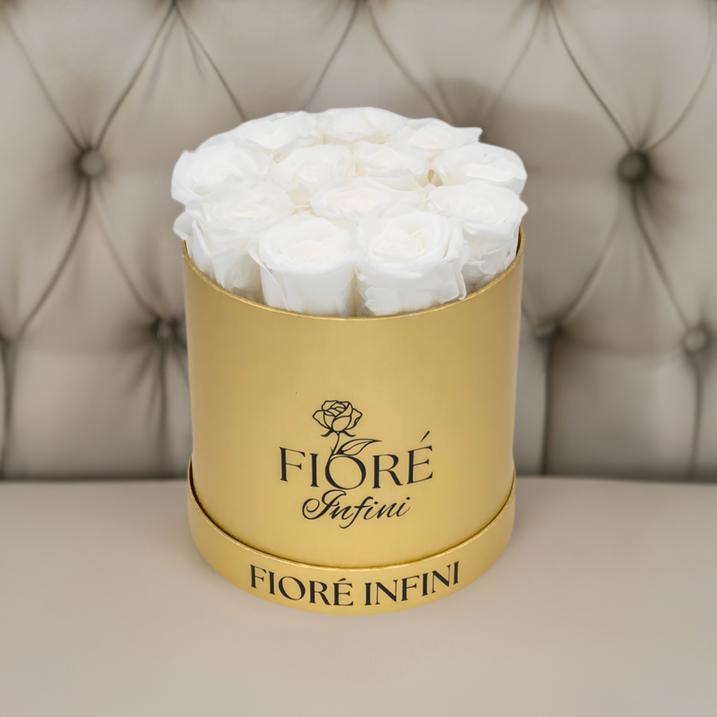 12 white forever roses in a gold foil box on a tan sofa