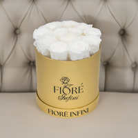 12 white forever roses in a gold foil box on a tan sofa